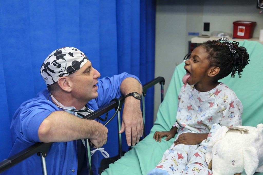 An African American child in a hospital bed being goofy as she sticks her tongue out at a white, male nurse