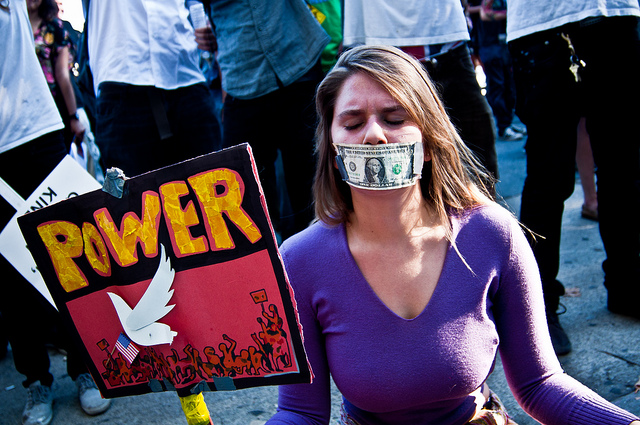 A woman with a dollar bill taped over her mouth, holding a sign that says