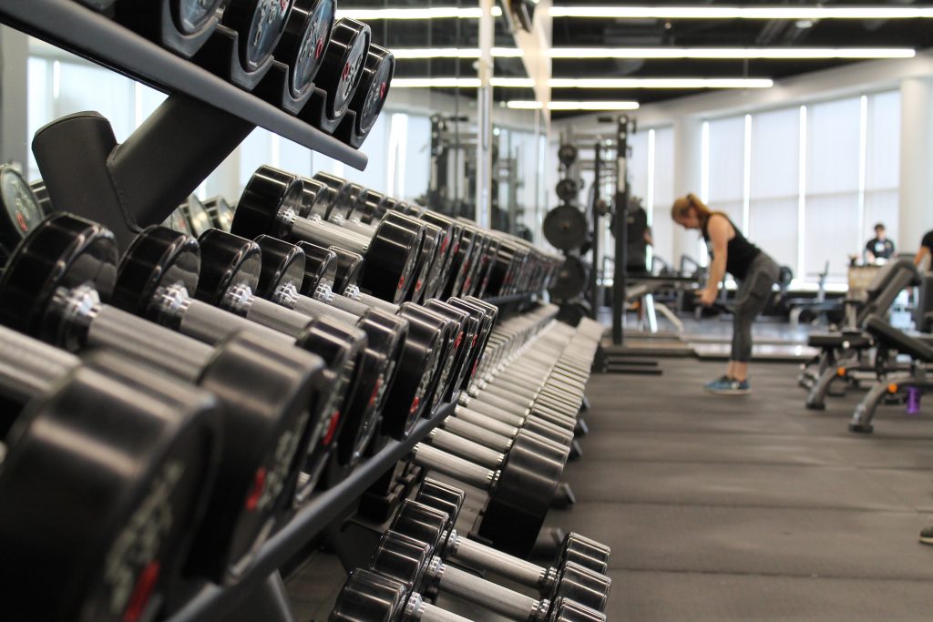 Room with rows of dumbbells with a woman lifting weights in the distance.