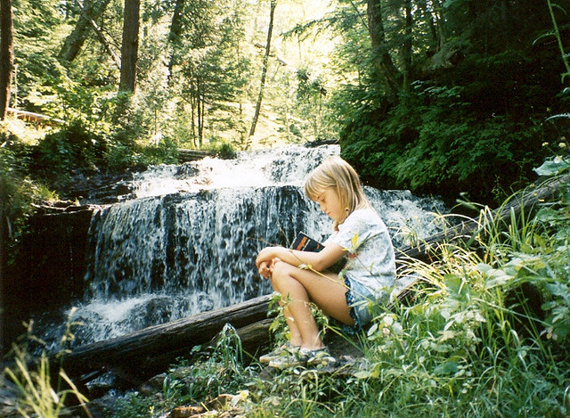A young girl reading a book on a rock near a peaceful waterfall
