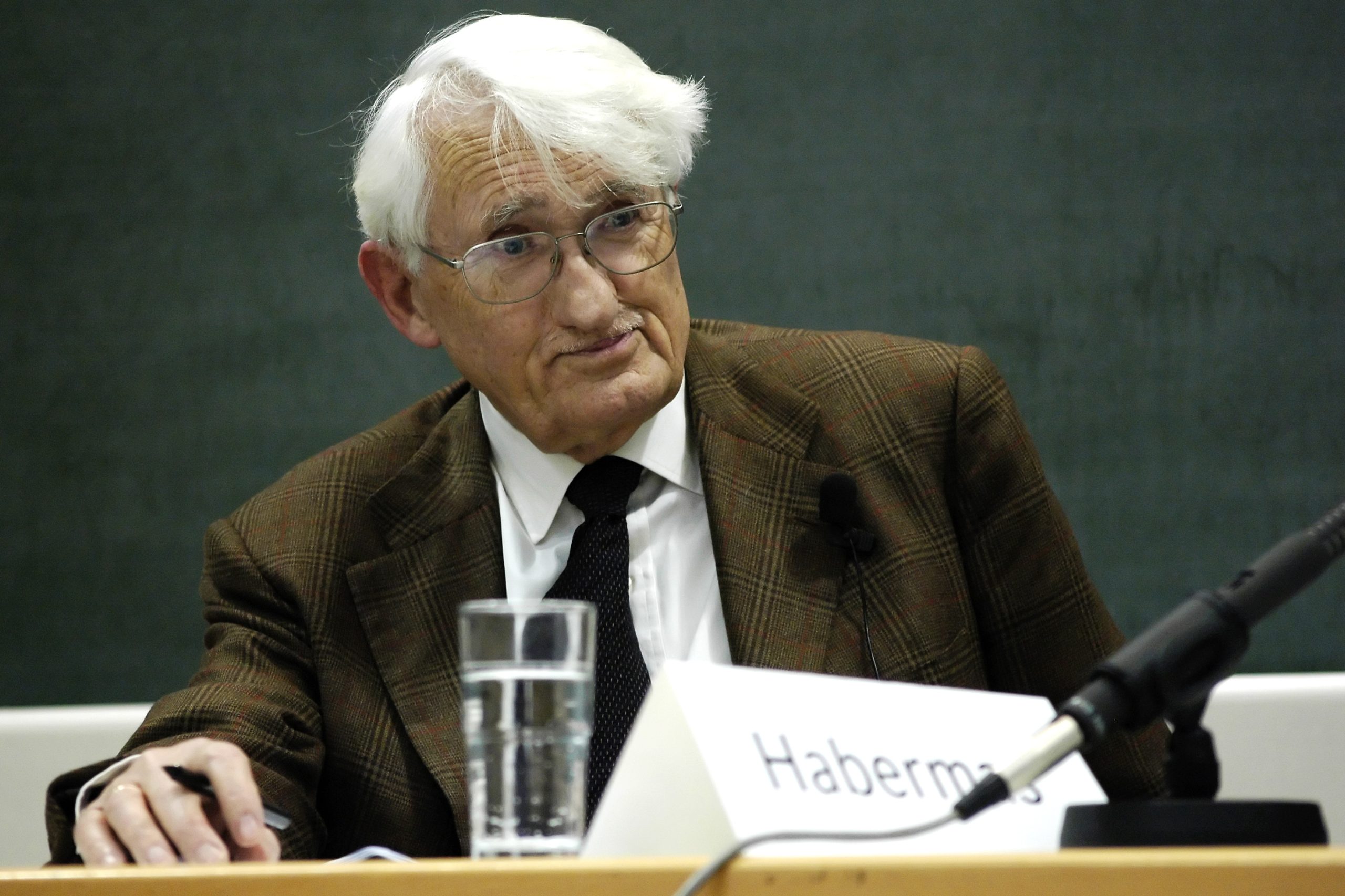 An image of German political philosopher Juergen Habermas, sitting at a table. A placard with Habermas's last name printed on it sits on the table in front of him.