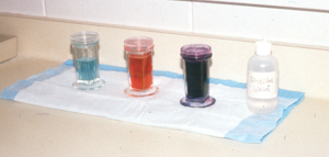 three containers of Diff Quick in blue, red, and purple and a bottle of distilled water on a paper towel